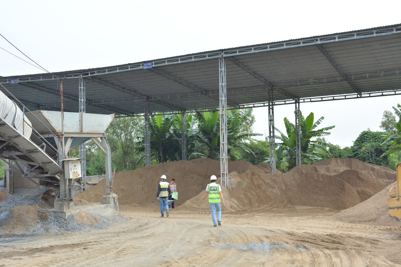 Industrial construction site with workers inspecting sand piles under metal roofing.